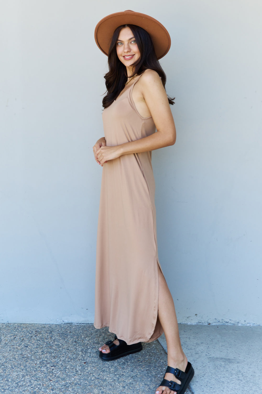 A woman stands against a plain background, wearing the Ninexis Good Energy Full Size Cami Side Slit Maxi Dress in Camel along with a wide-brimmed hat. She has one hand on her hip and is smiling.