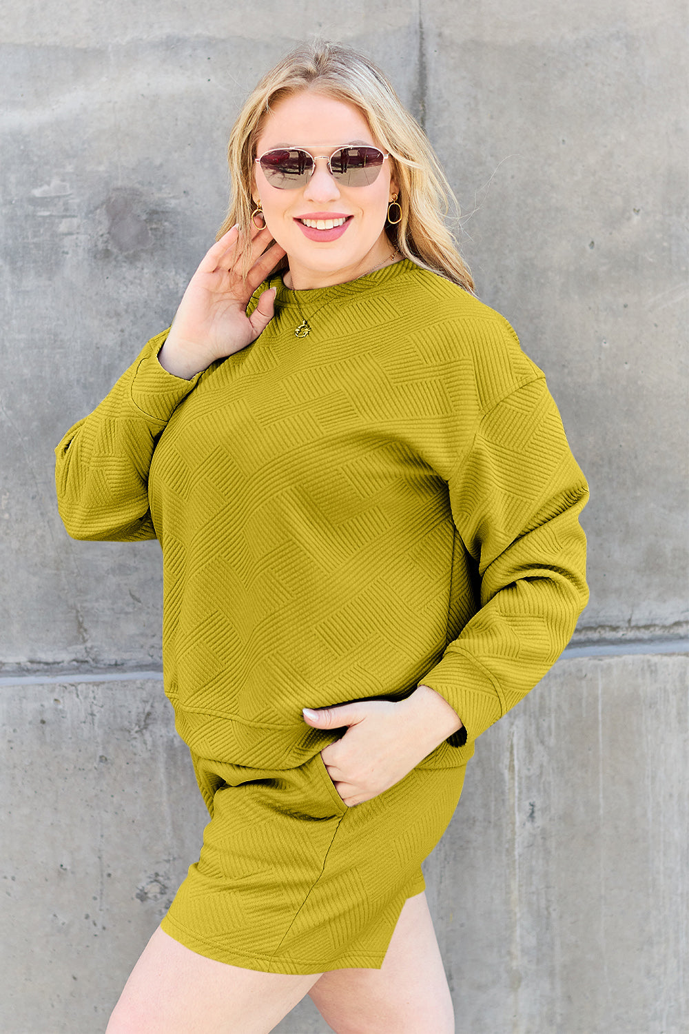 A woman decked out in the Double Take Full Size Texture Long Sleeve Top and Drawstring Shorts Set, featuring a brown textured two-piece with a long-sleeve top and matching pocketed shorts, smiles at the camera.