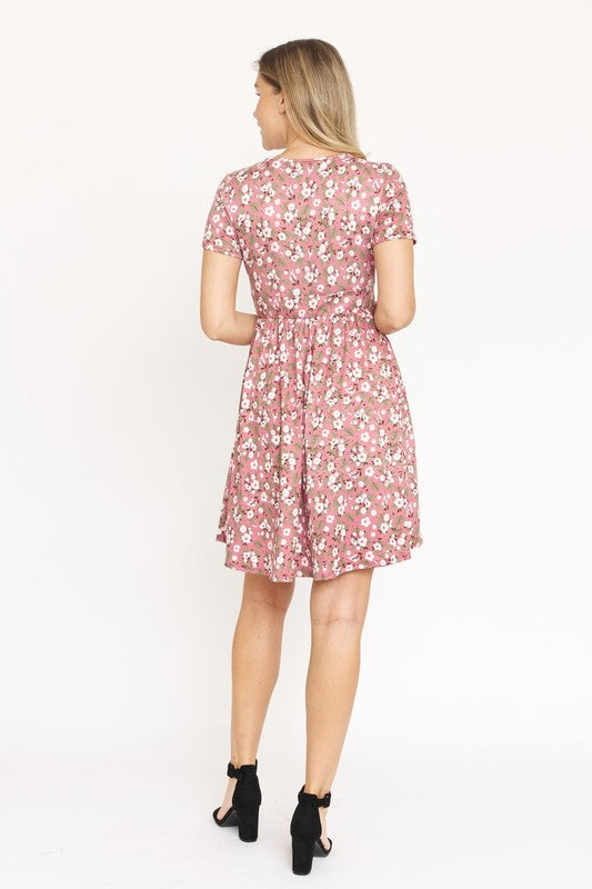 A woman wearing a short-sleeved, V-neck, Ditsy Surplice Fit and Flare Midi Dress with a dark floral background, crafted from luxurious Polyester Spandex and proudly Made in the United States, poses against a white backdrop, smiling as she softly holds the sides of her dress.