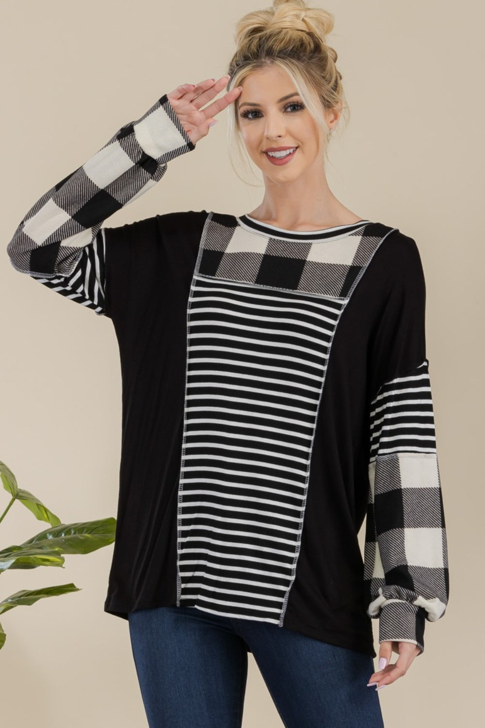 A person wearing a Celeste Full Size Color Block Exposed Seam T-Shirt, featuring black and white plaid with reverse stitching and long sleeves, poses while smiling in front of a neutral background with a plant.