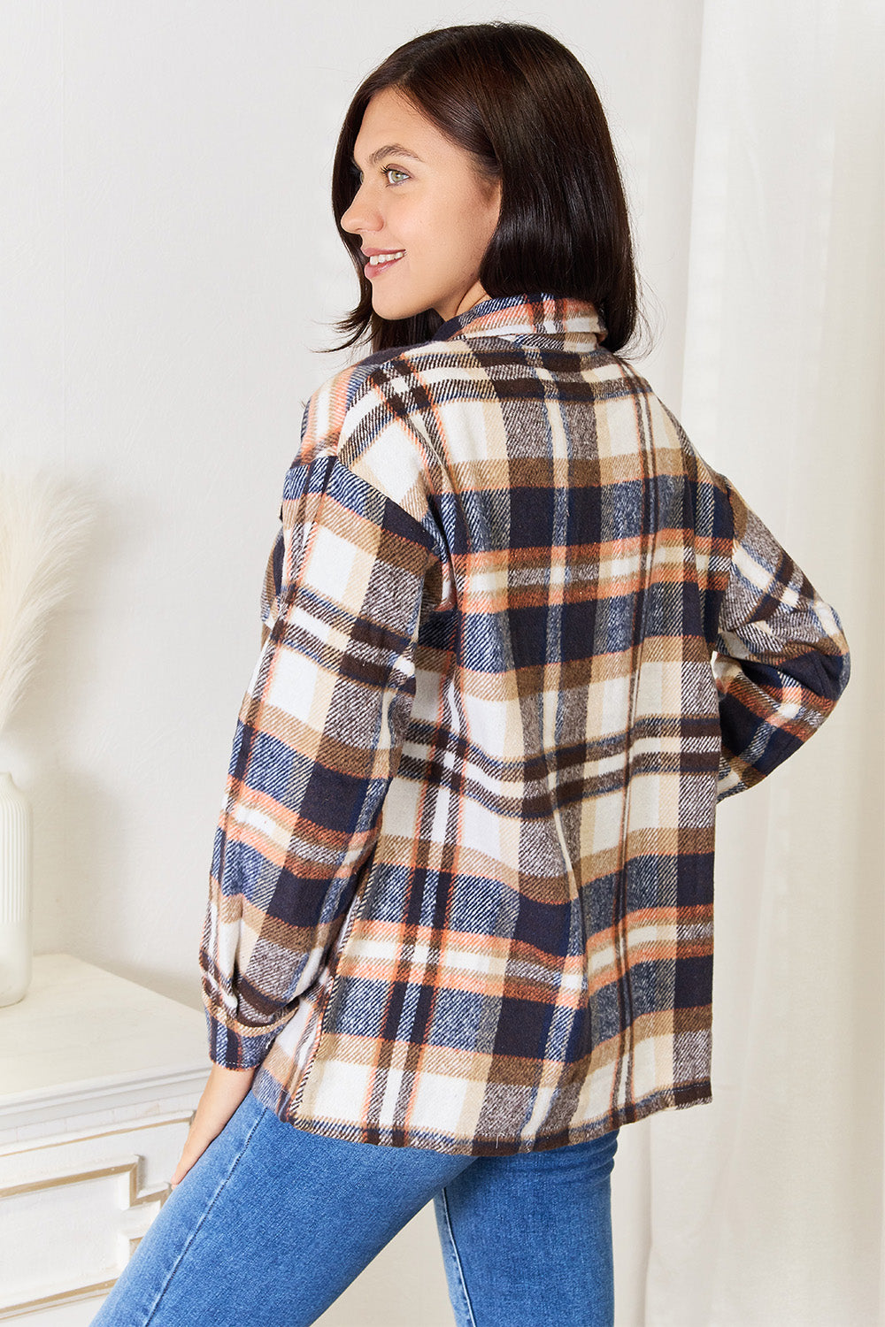 A person with long braided hair wearing the Double Take Plaid Button Front Shirt Jacket with Breast Pockets over a white top and blue jeans stands with a hand in their pocket, posing in an indoor setting. The layering of the jacket adds an effortless touch to the outfit.