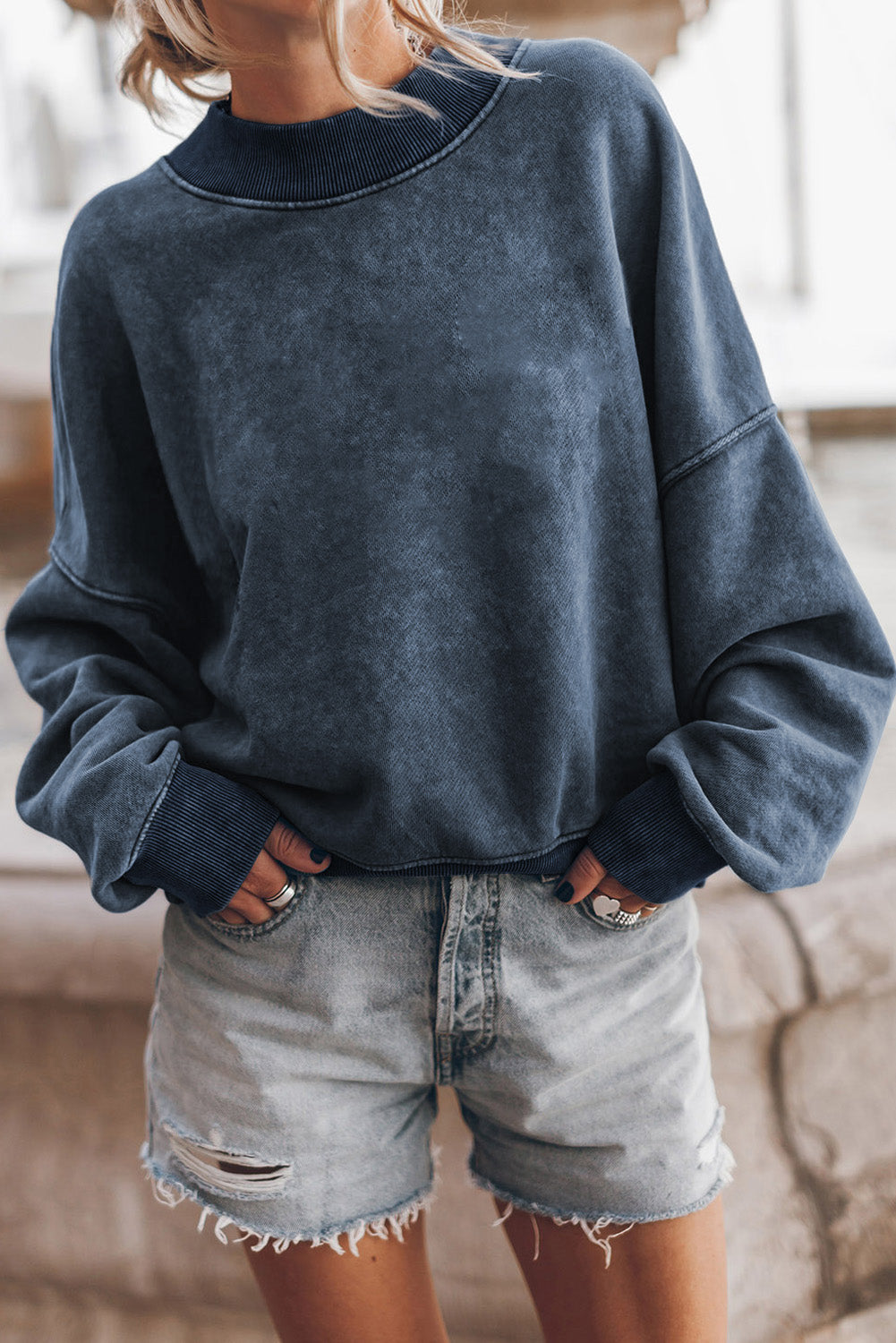 A person with long blonde hair is wearing a sky blue drop shoulder crew neck pullover sweatshirt and light denim shorts, standing outdoors with their back to the camera.