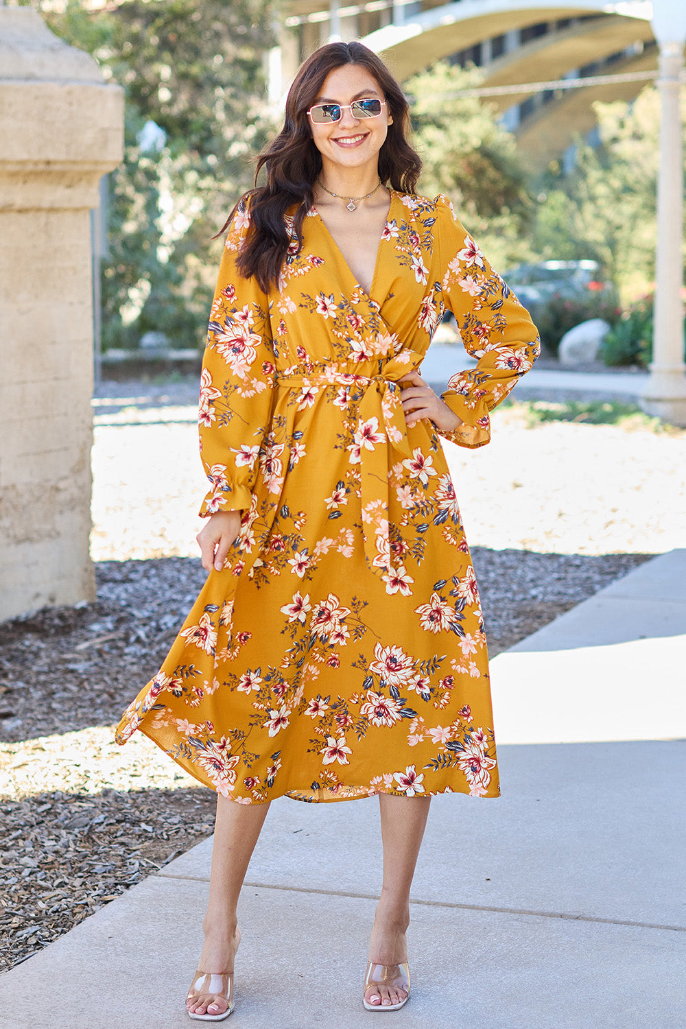 A woman wearing the Double Take Full Size Floral Tie Back Flounce Sleeve Dress in yellow, along with sunglasses, is walking on a paved path outdoors. She is smiling and has one hand on her hip.