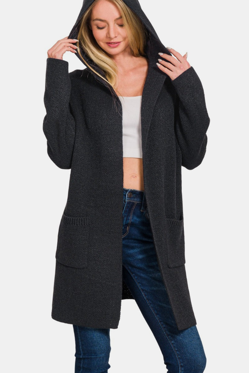 A person stands with hands gripping the hood of a Zenana Hooded Open Front Sweater Cardigan, effortlessly paired with a white crop top and jeans—the perfect cozy layering piece for any casual outing.