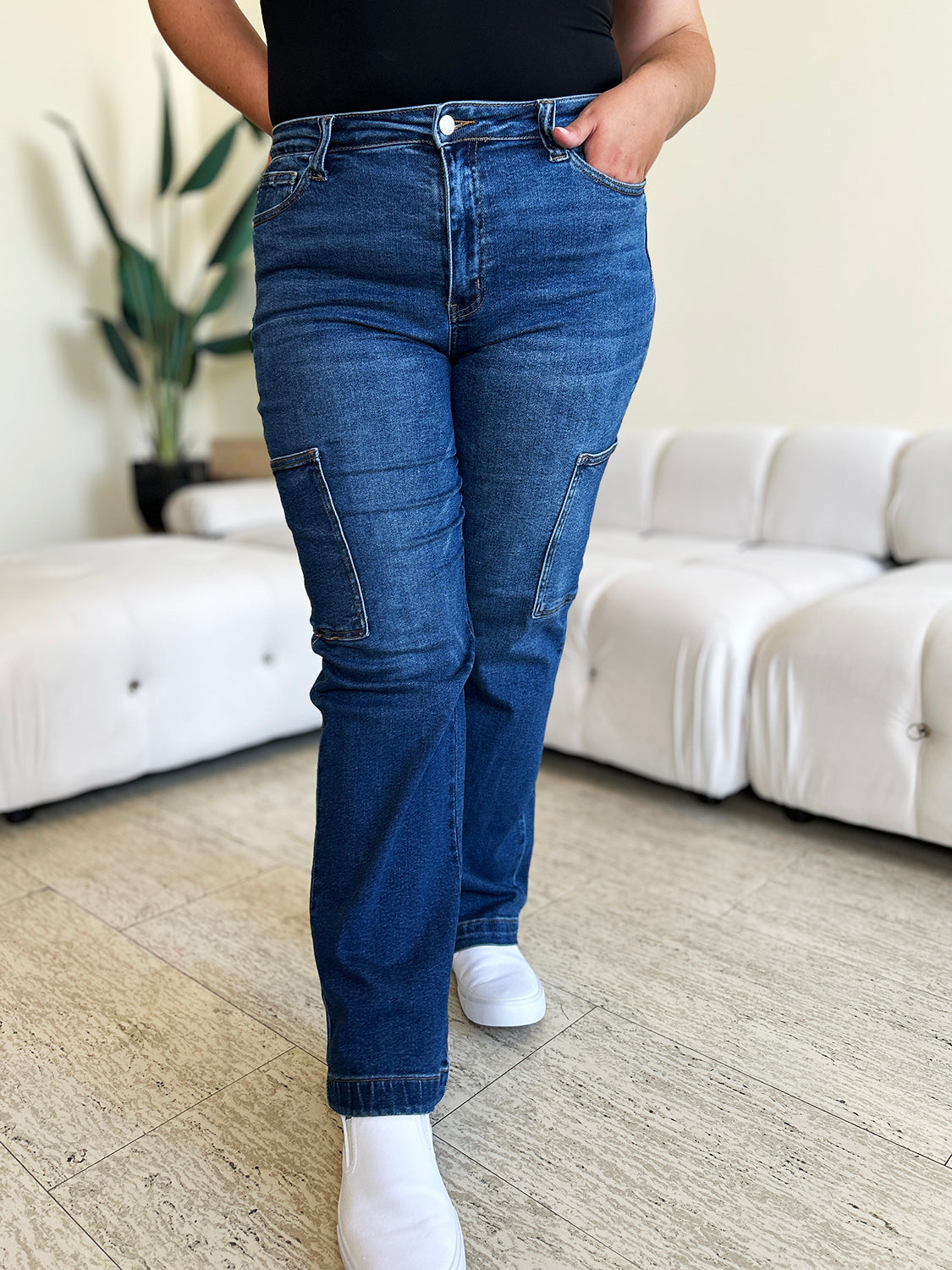 A person wearing Judy Blue Full Size High Waist Straight Cargo Jeans made of durable denim fabric and a white top walks on a wooden floor, with a white sofa and green plant in the background.