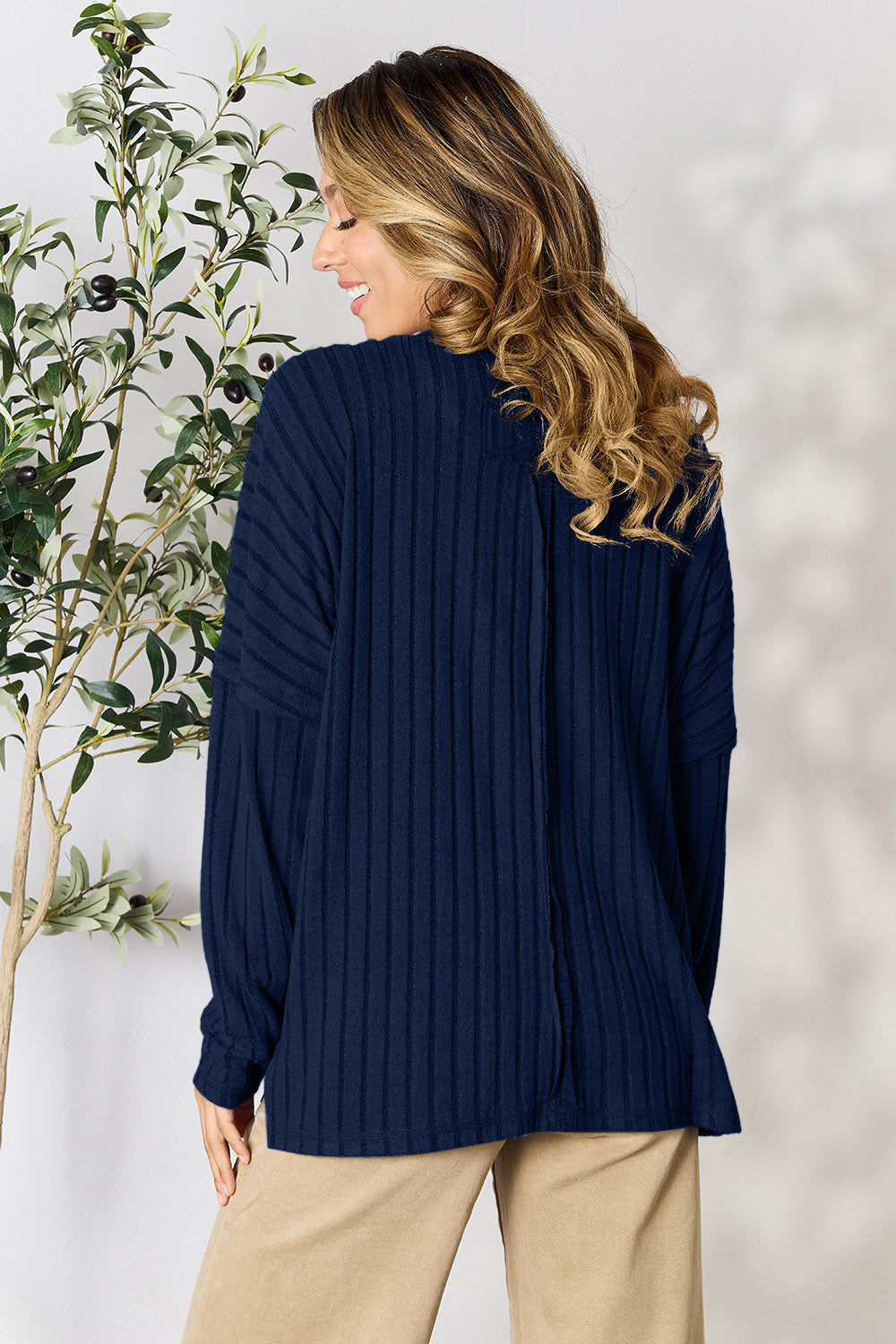 A person wearing the Basic Bae Full Size Ribbed Round Neck Slit Knit Top in blue and beige pants stands and smiles in front of a leafy plant.