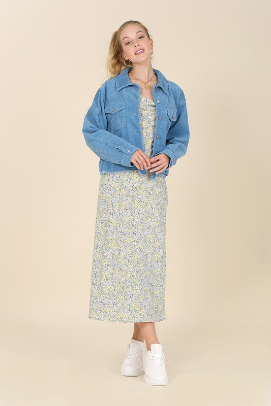 A person with long hair wears a blue frayed corduroy jacket over a floral dress, seated on a light-colored background.