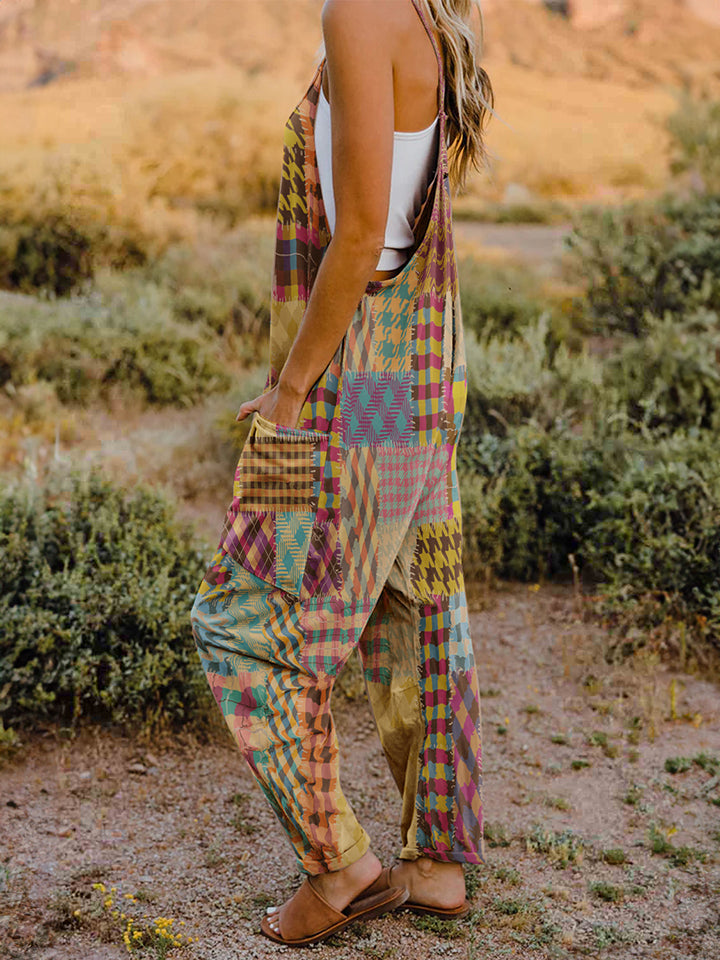 A woman stands outdoors in front of a rocky landscape, wearing the Full Size Printed V-Neck Sleeveless Jumpsuit and a brown brimmed hat. She is smiling and holding the brim of her hat with one hand, exuding the carefree vibe of this vibrant and colorful jumpsuit.