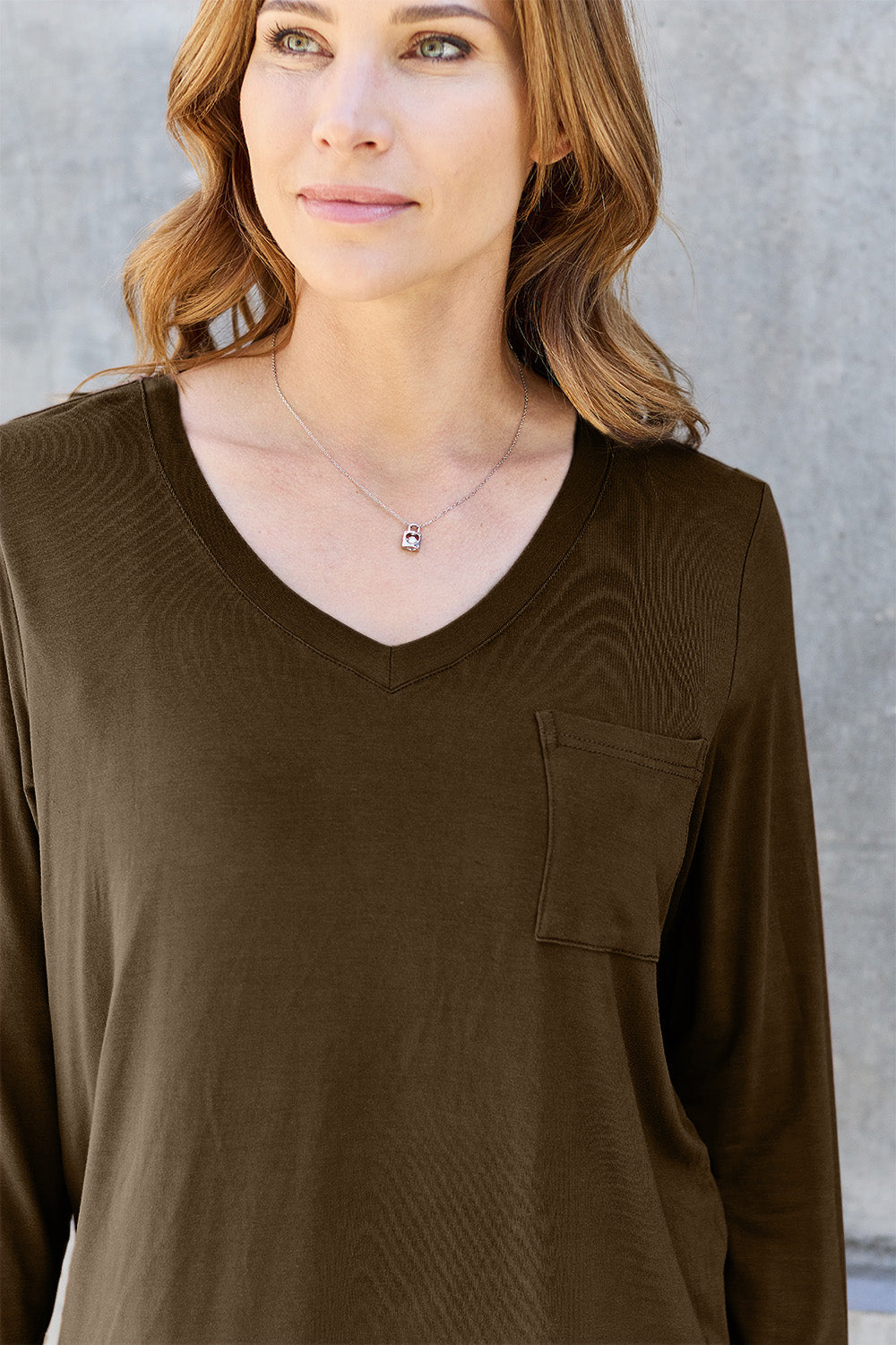 A woman with light brown hair stands against a concrete wall, wearing the Basic Bae Full Size V-Neck Long Sleeve Top in olive green and blue jeans, with a white backpack on her shoulder. Her outfit exudes basic style and is made from polyester, ensuring it’s both comfortable and suitable for machine wash cold. She is looking at the camera.