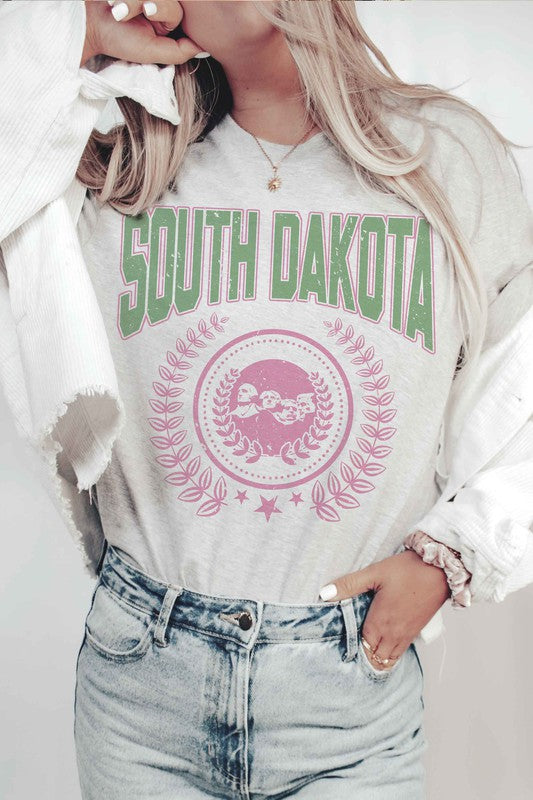 A person wearing a white SOUTH DAKOTA Graphic Tee, adorned with green and pink lettering, along with matching white shorts, stands indoors. The unisex sizing 100% cotton tee adds a comfortable yet stylish touch to their outfit.