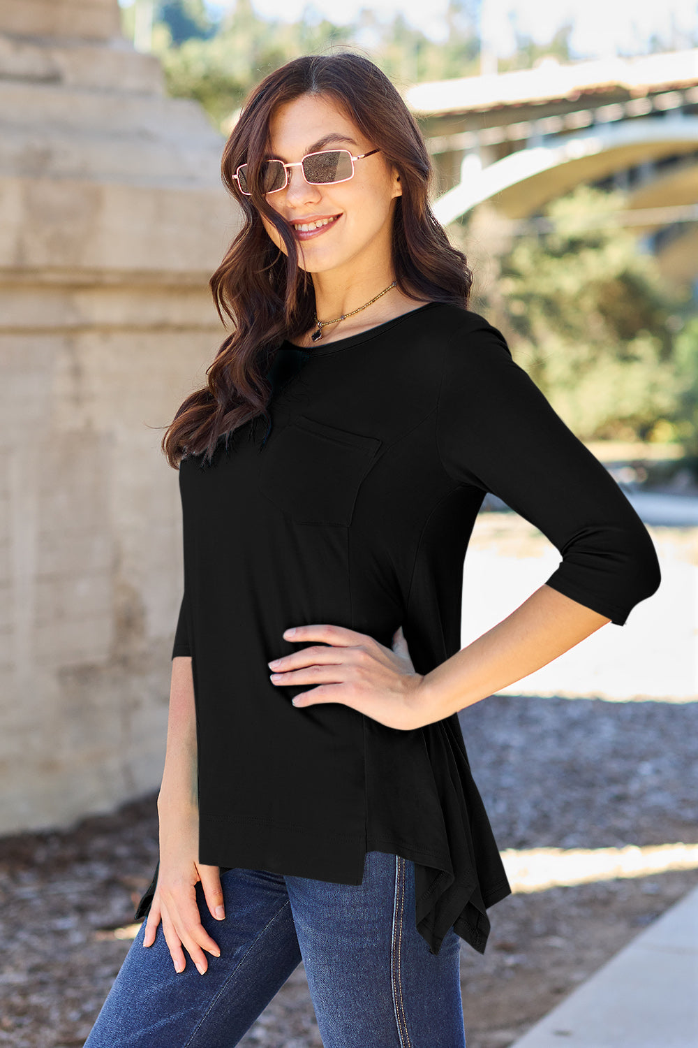 A woman stands outdoors wearing sunglasses, a Basic Bae Full Size Round Neck Pocketed T-Shirt with an asymmetrical hem and chest pocket made of rayon spandex, and blue jeans. She is smiling and has one hand in her pocket. Her basic style radiates effortless charm.