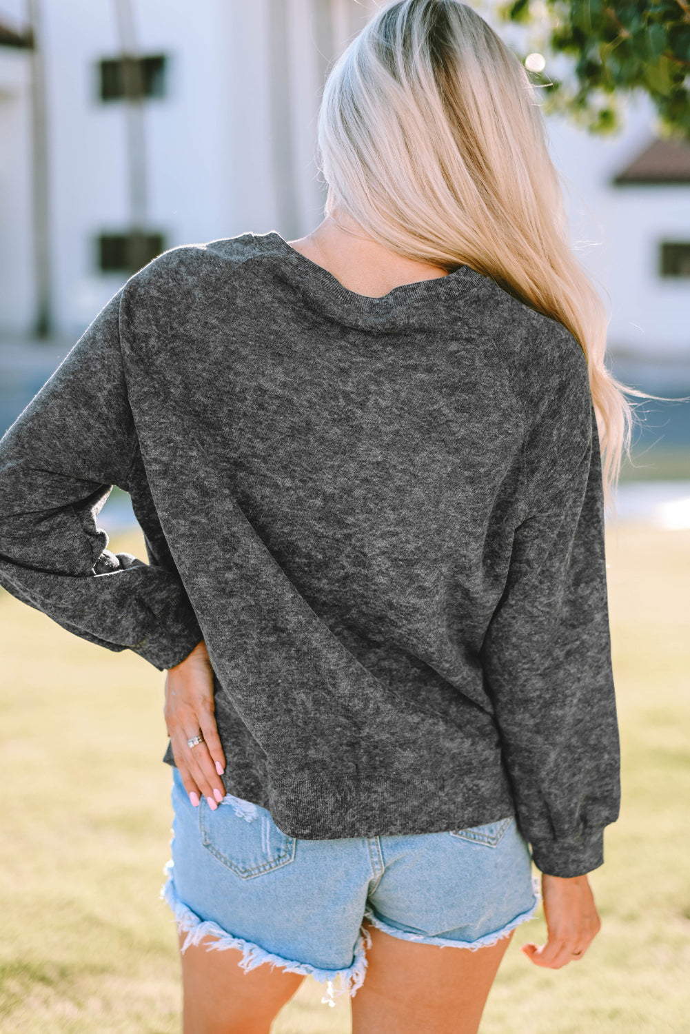 A blonde woman wearing a Gray Coors Banquet RODEO Graphic Mineral Washed Sweatshirt and light blue denim shorts, showcasing casual outdoor wear, stands outdoors with her back to the camera.