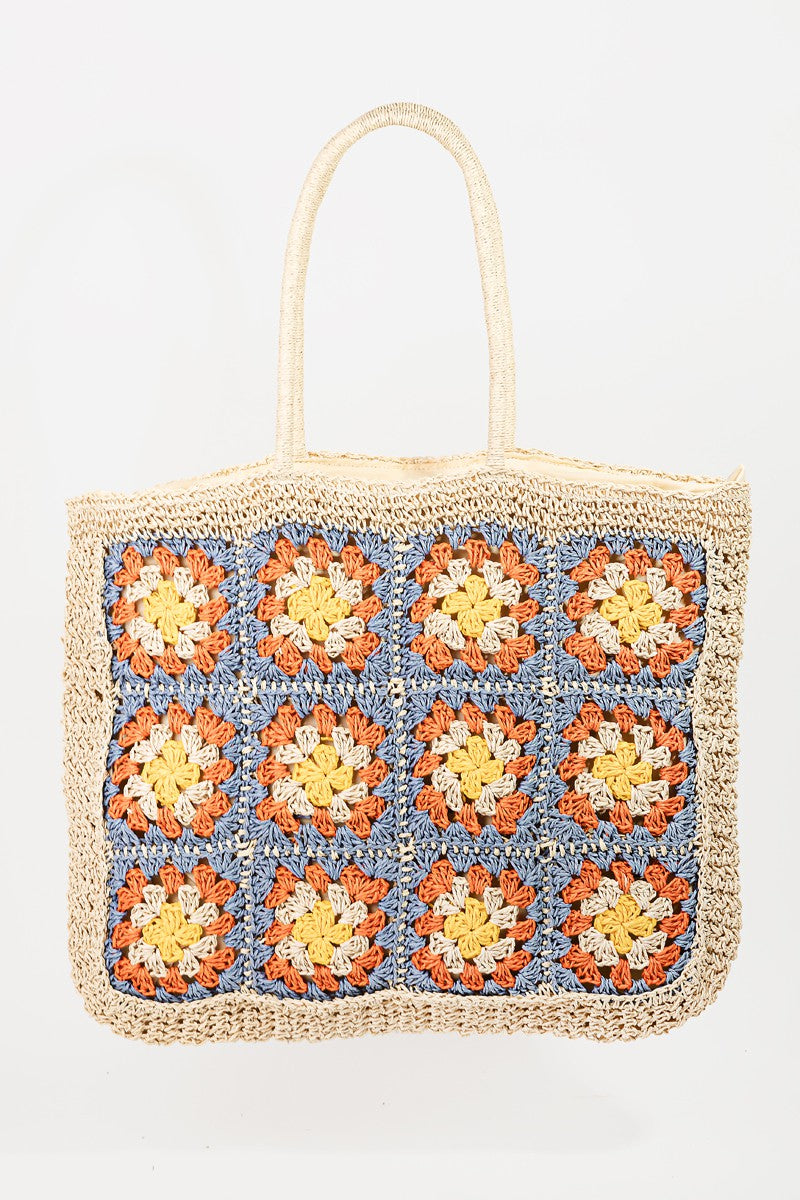 The Fame Flower Braided Tote Bag is a crocheted accessory with bohemian flair, featuring a grid of colorful flower patterns on the front. It showcases squares in orange, yellow, and blue on a natural fiber background.