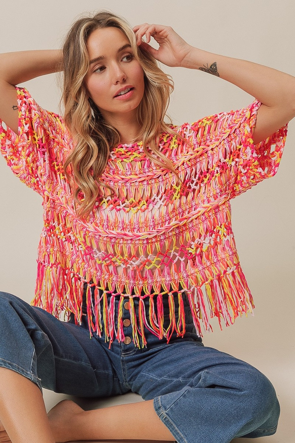 A person with long hair wears the BiBi Openwork Fringed Knit Cover Up, exuding bohemian flair, paired with jeans. They sit with one hand raised and the other resting on their lap against a plain background.