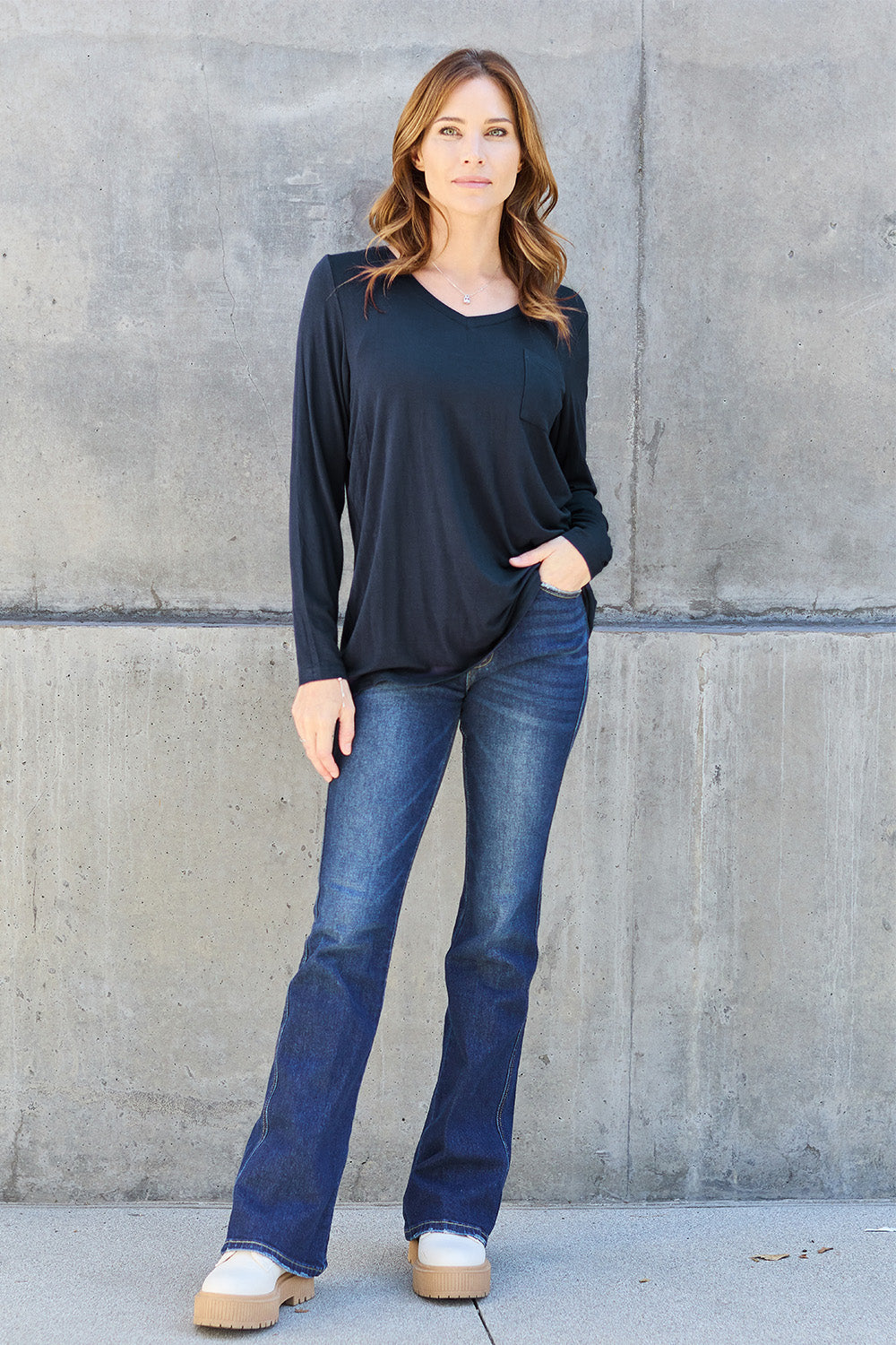 A woman with light brown hair stands against a concrete wall, wearing the Basic Bae Full Size V-Neck Long Sleeve Top in olive green and blue jeans, with a white backpack on her shoulder. Her outfit exudes basic style and is made from polyester, ensuring it’s both comfortable and suitable for machine wash cold. She is looking at the camera.