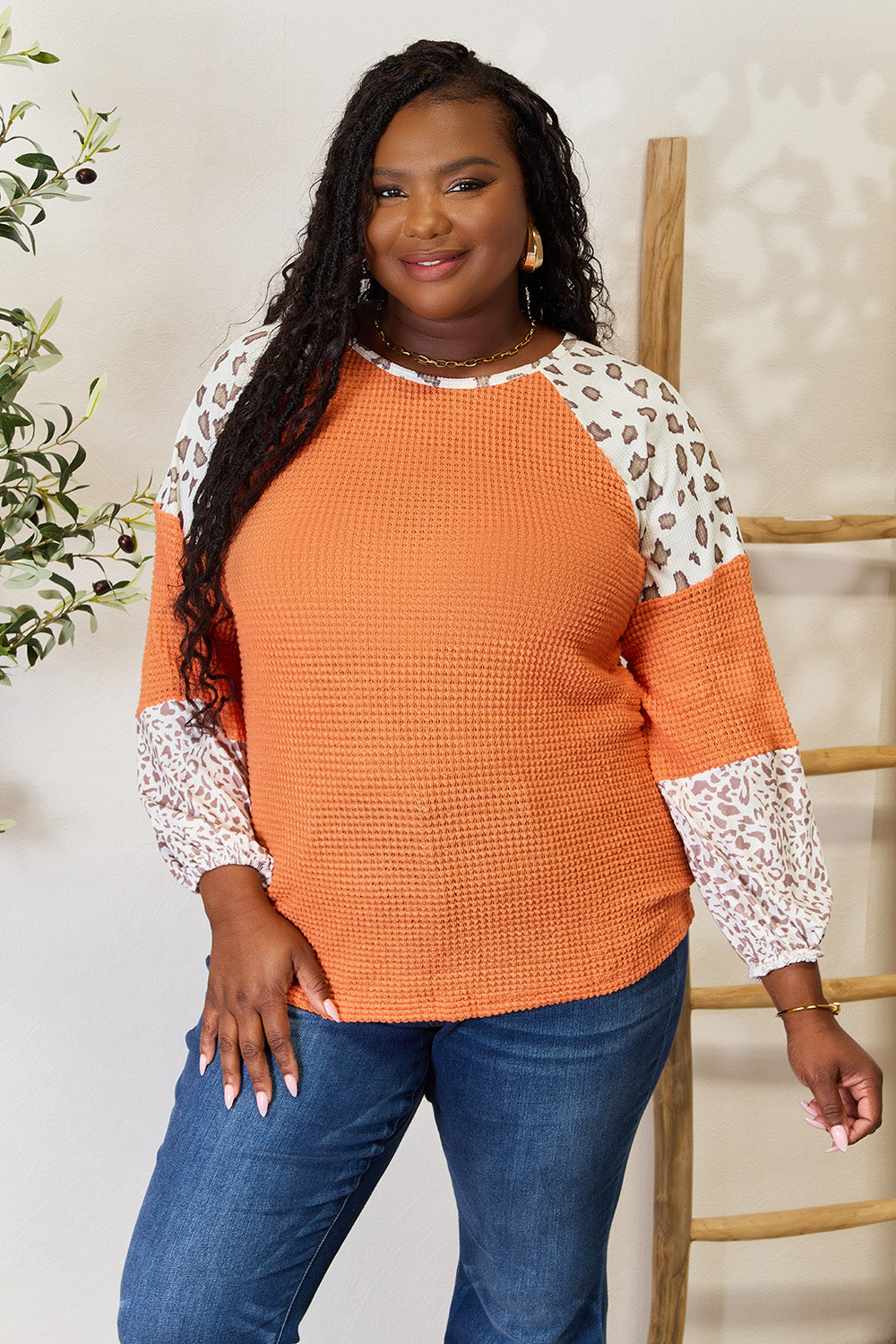 A woman poses, smiling, wearing the trendy Leopard Waffle-Knit Long Sleeve Blouse paired with blue jeans, standing near a wooden ladder and a plant.