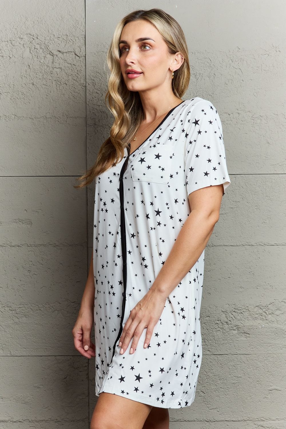 A woman with long hair wearing a MOON NITE Quilted Quivers Button Down Sleepwear Dress, adorned with black stars, stands against a textured gray wall, posing with one hand on her hip.