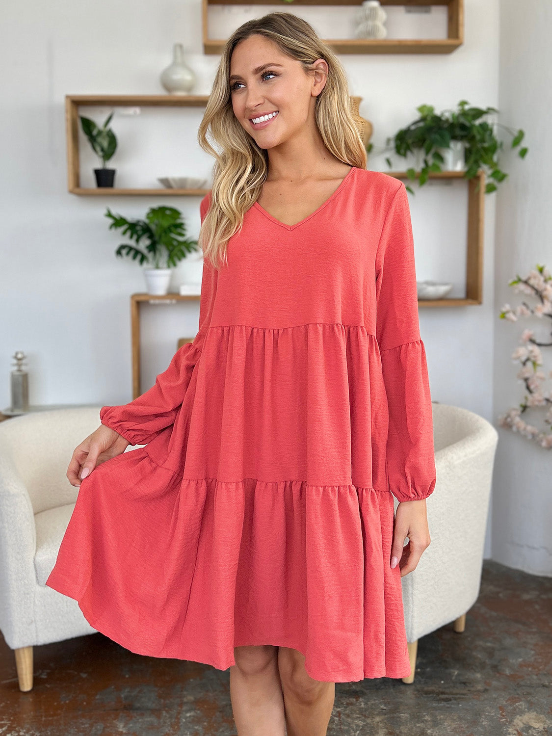 A woman stands wearing the Double Take Full Size V-Neck Balloon Sleeve Tiered Dress with Pockets in pink. Crafted from high stretch yarn, this dress combines comfort and style effortlessly. She has long, wavy hair and accessorizes with hoop earrings and a necklace. This imported garment is easy to care for – simply machine wash cold to keep it looking fresh.