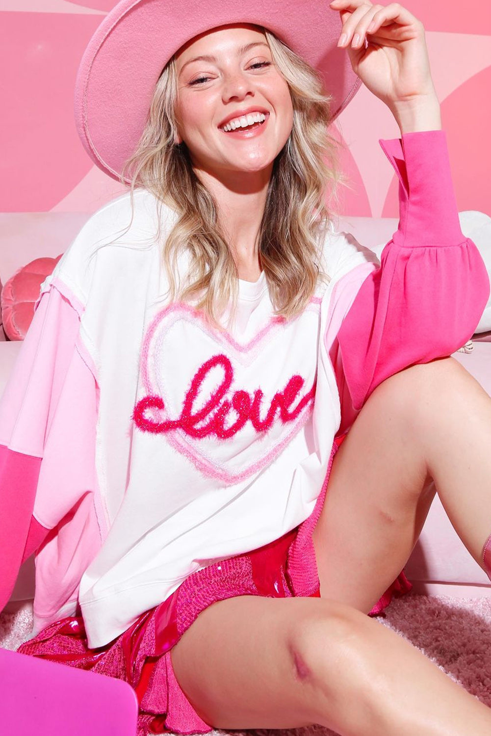 A woman with long blonde hair is smiling and posing indoors. She is wearing a White Tinsel Love Heart Graphic Color Block Sleeve Sweatshirt with pink shorts. There are framed photos and a table in the background.