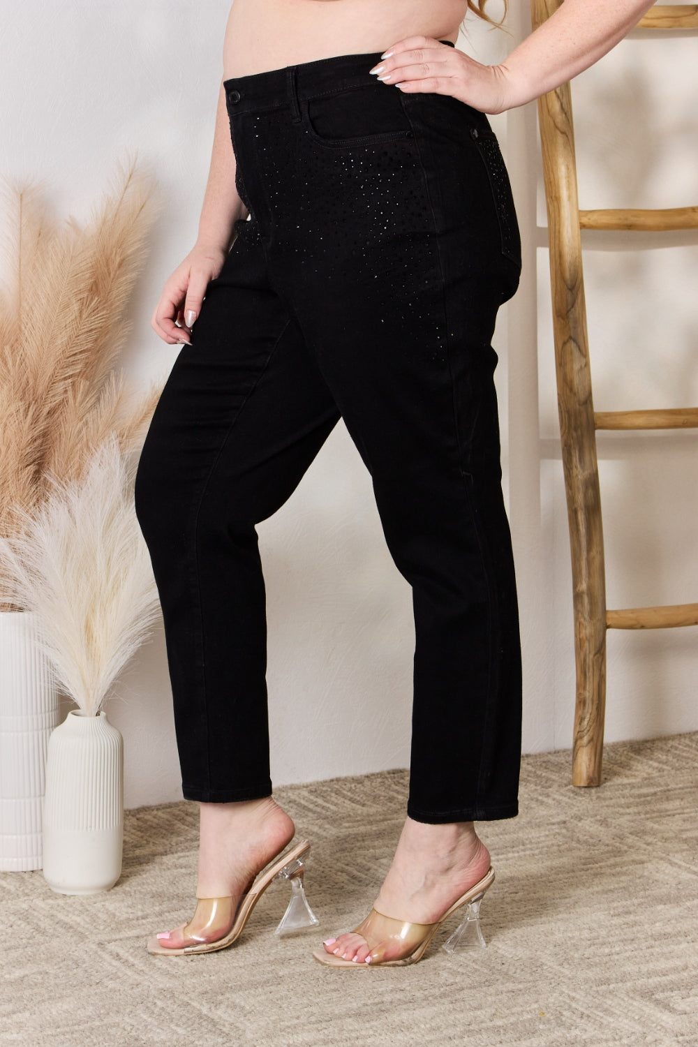 A person wearing slightly stretchy, black Judy Blue Full Size Rhinestone Embellished Slim Jeans and black shoes stands on a textured floor next to a white vase with brown and white ornamental grasses and a wooden ladder.