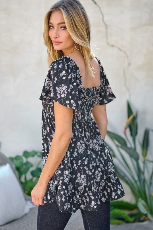 A woman stands outdoors donning a Floral Printed V-Neck Ruffle Top paired with jeans, set against a neutral background, with plants in the foreground, highlighting her fashion-forward wardrobe.