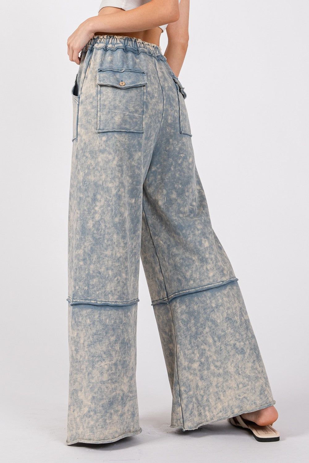 A person is wearing the SAGE + FIG Mineral Washed Terry Wide Leg Pants, which are high-waisted and light blue with a tie-dye appearance. Made from breathable cotton, they feature a drawstring elastic waistband and large front pockets, paired with beige sandals.