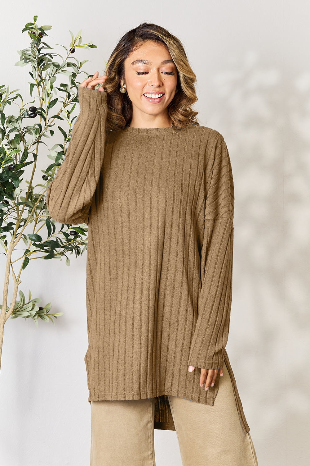 A person with shoulder-length hair smiles while wearing a beige ribbed sweater called the Basic Bae Full Size Ribbed Round Neck Long Sleeve Slit Top, along with matching beige pants, showcasing a basic style. They stand next to a green plant.