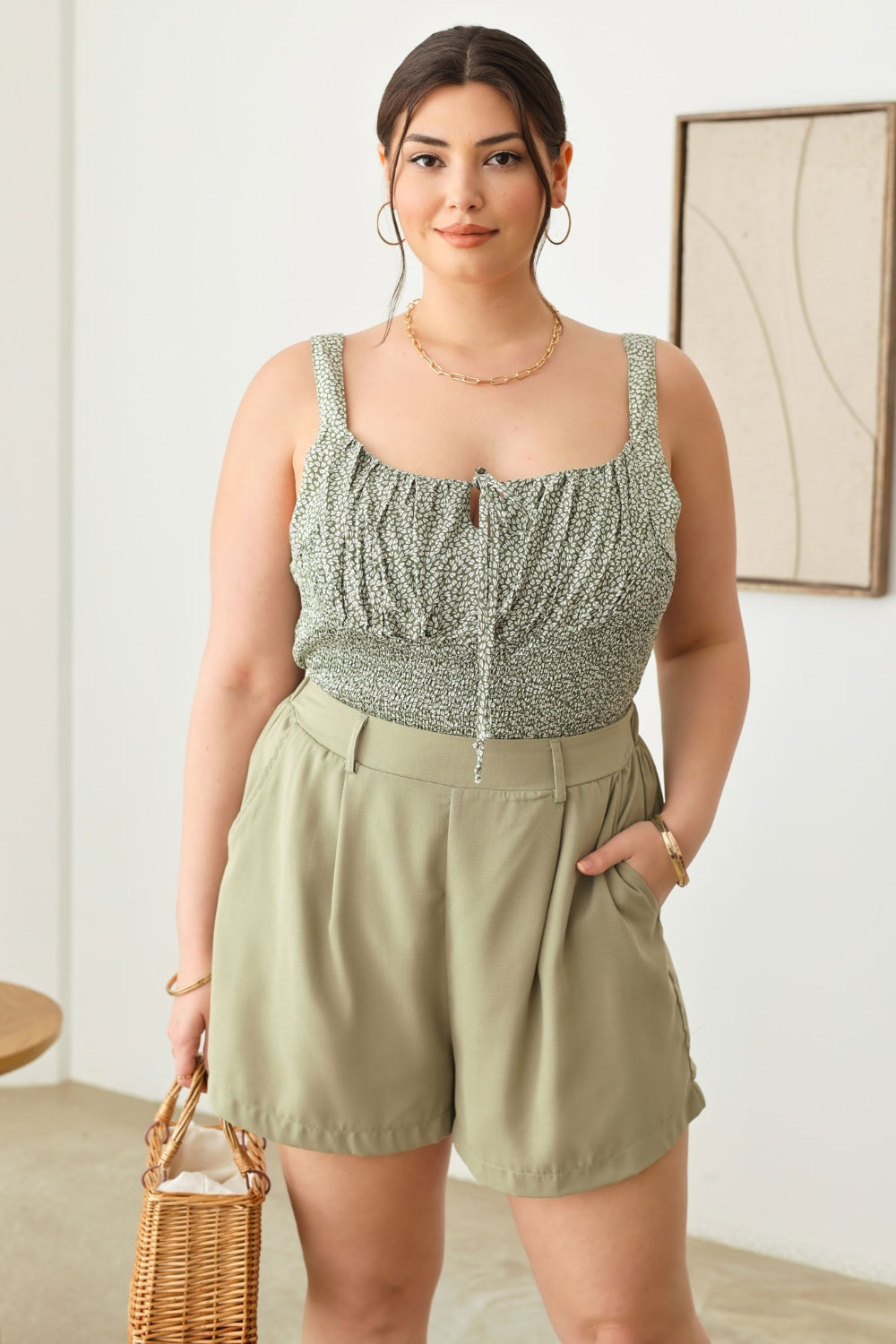 A woman in a green sleeveless top and the Zenobia Plus Size Half Elastic Waist Shorts, featuring convenient pockets, holds a woven handbag while standing indoors.