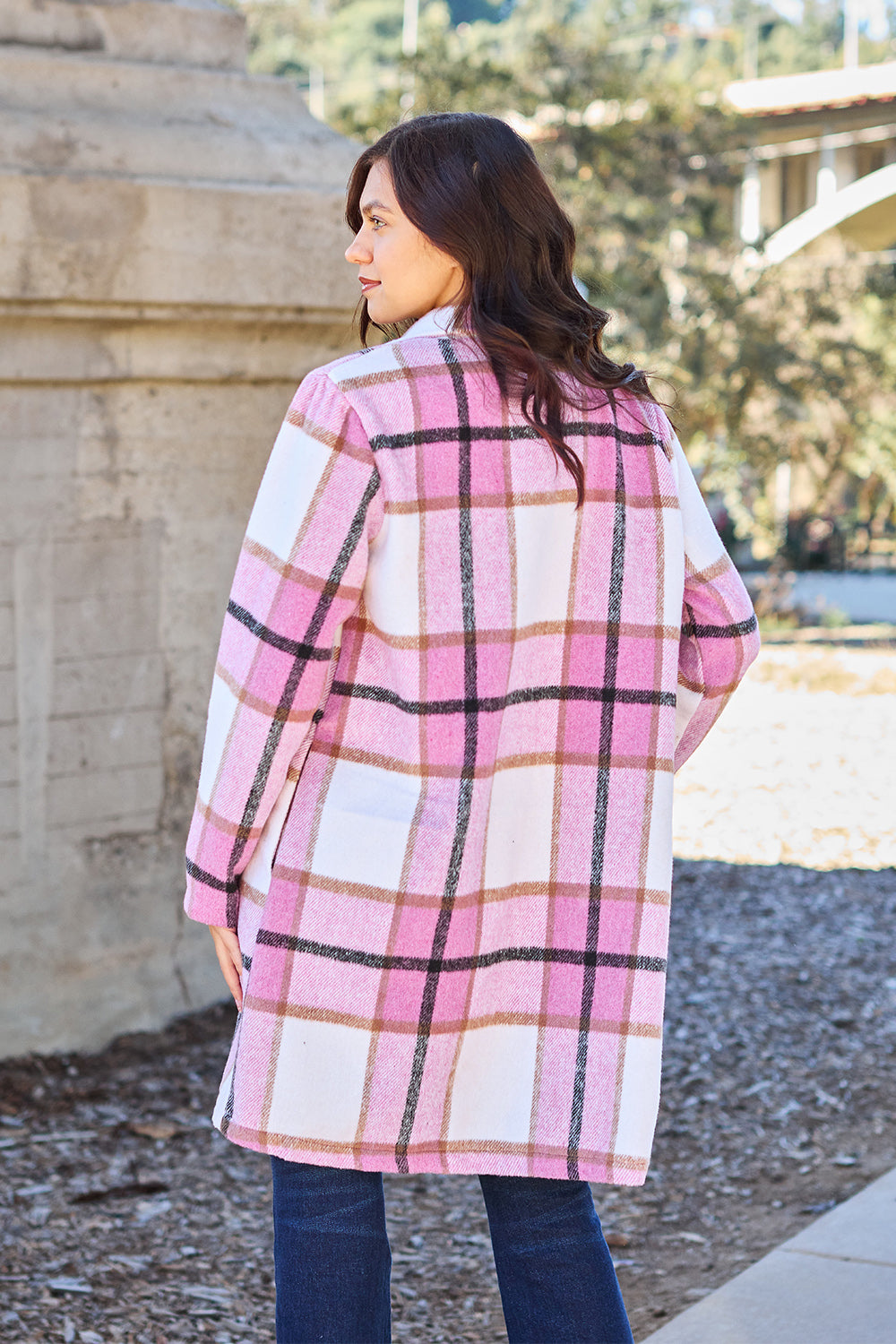 A woman stands on a sidewalk wearing the Double Take Full Size Plaid Button Up Lapel Collar Coat in pink and white over a white top and blue jeans, with her right hand lightly touching her hair. The polyester fabric of the coat makes it durable and perfect for an easy machine wash cold.