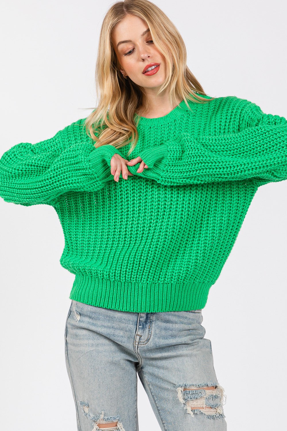 A person wearing a SAGE + FIG round neck drop shoulder sweater, ripped light blue jeans, and a green hat poses against a white background.