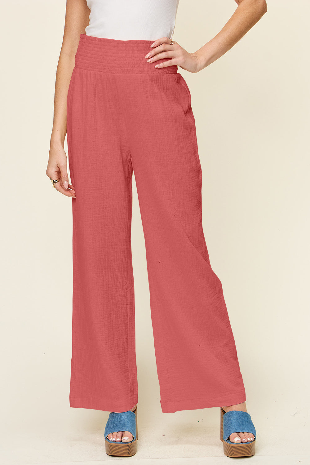 A person wearing Double Take Full Size Texture Smocked Waist Wide Leg Pants in black and a white 100% cotton top is standing with one hand on their hip. They are also wearing blue open-toe sandals.