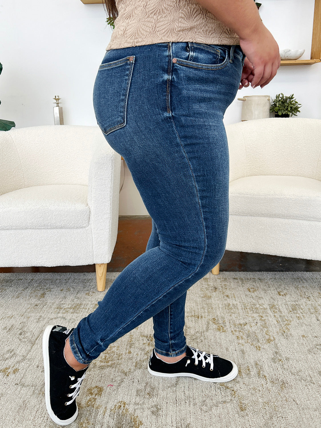 A person stands indoors wearing Judy Blue Full Size Classic Handsand Skinny Jeans, a beige top, and black sneakers with white laces. Their right hand is in their pocket. A partial view of two white chairs is visible in the background. The jeans feature a hand-sanded finish for a flattering fit.