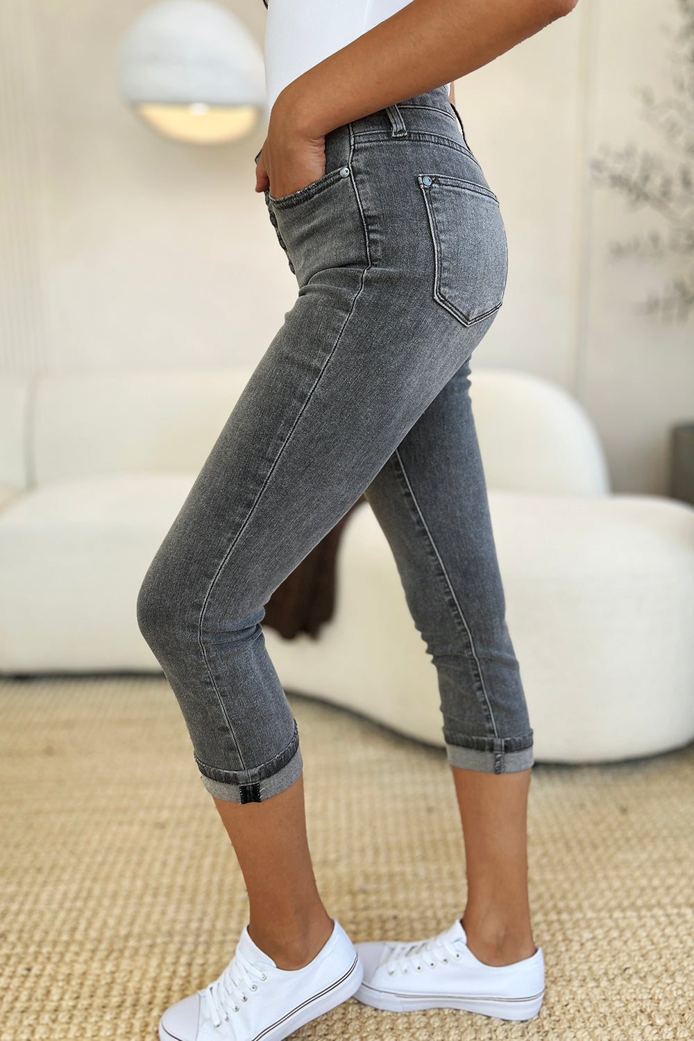 A person wearing Judy Blue Full Size Button Fly High Waist Cuffed Capris in gray, paired with a white top and white sneakers, stands on a beige carpeted floor.