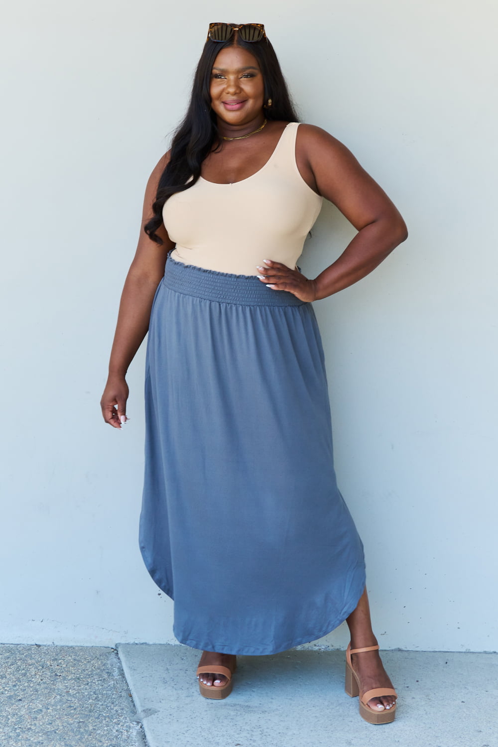 A person wearing a white sleeveless top and the Doublju Comfort Princess Full Size High Waist Scoop Hem Maxi Skirt in Charcoal stands against a plain wall. They are also wearing black sandals.