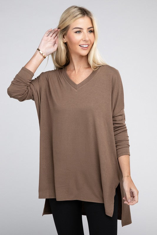 A woman with long, blonde hair is wearing a loose-fitting, brown Dolman Long Sleeve V-Neck Side Slit Hi-Low Hem Top and black pants. She poses with one hand on her hip and a neutral facial expression against a light grey background.