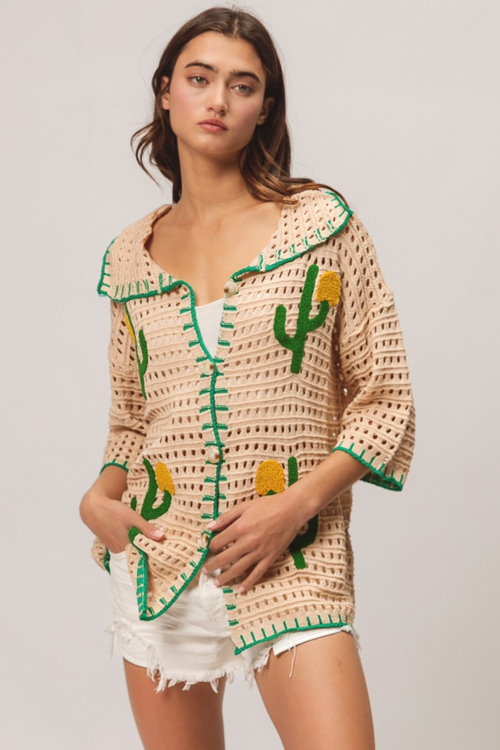 A woman wearing the BiBi Edge Stitched Cactus Patch Sweater Cardigan, featuring beige fabric with green cactus designs, layered over a white top and paired with white frayed shorts, stands against a plain background.