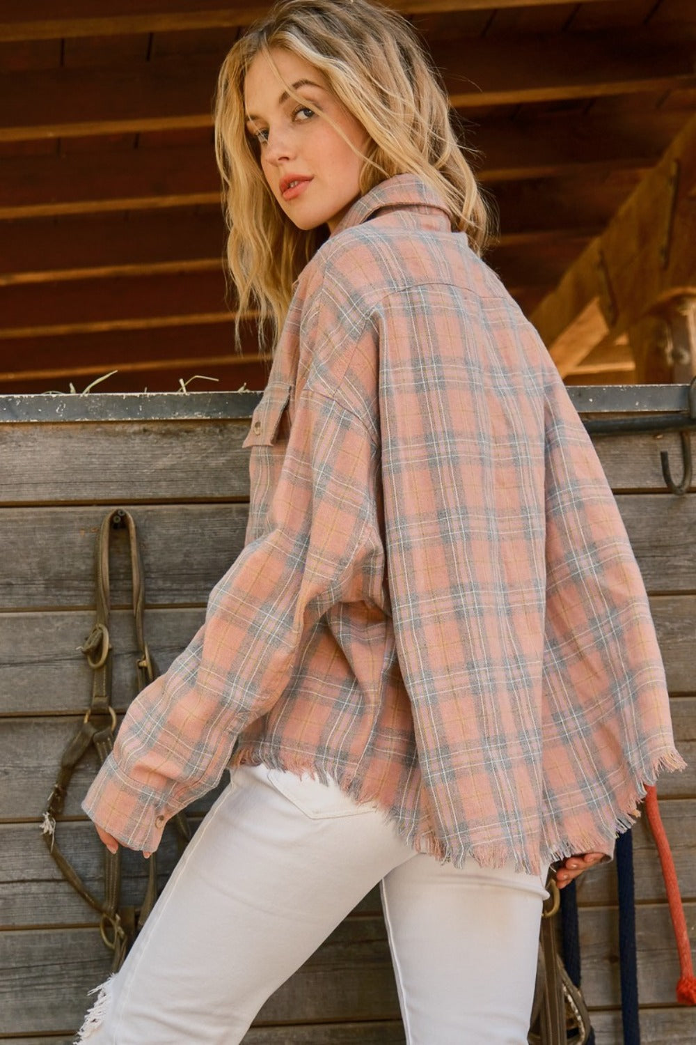 A person wearing the And The Why Full Size Plaid Button Up Raw Hem Shirt and white pants is standing in front of a wooden structure, showcasing raw edge detailing, with bridles hanging beside them.