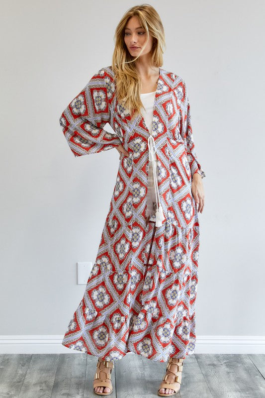 A person wearing a Printed Long Sleeve Loose Kimono with a bohemian blue and pink print over a white outfit stands in a room with a gray floor and white wall.