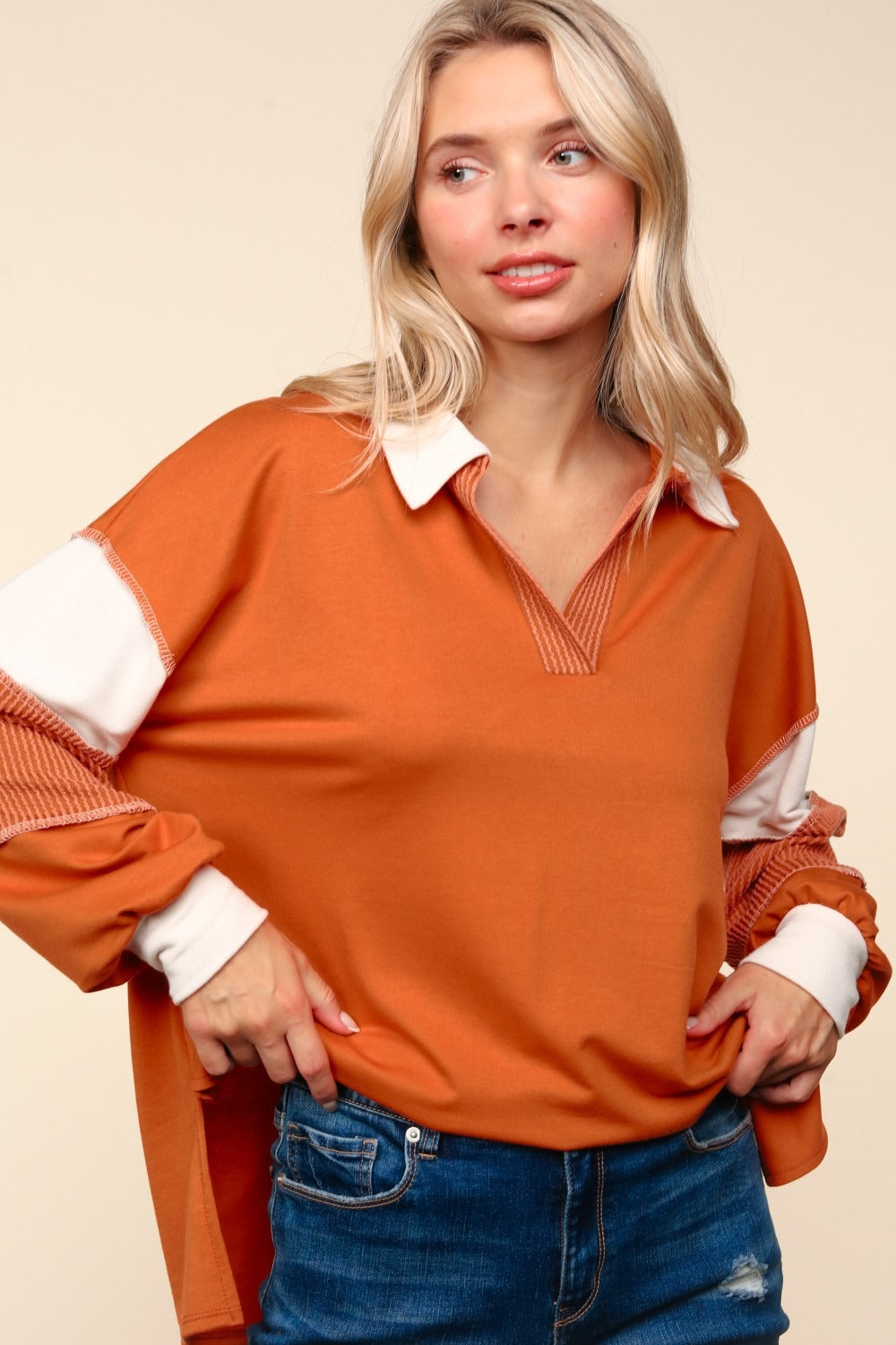 A person with blonde hair is wearing the Haptics Color Block Exposed Seam Long Sleeve Top in orange with white and orange sleeves, paired with blue jeans, standing against a beige background.