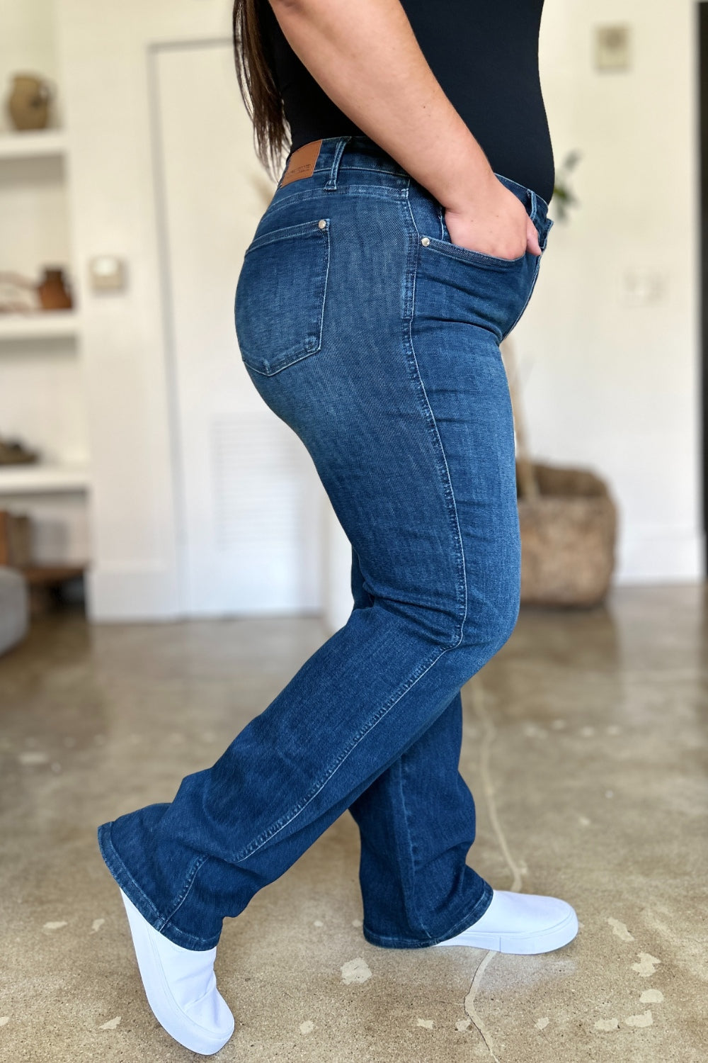 A person wearing a black top and Judy Blue Full Size Tummy Control Straight Jeans stands indoors with one hand on their hip. Their shoes are black with white soles. A plant and shelves are in the background.