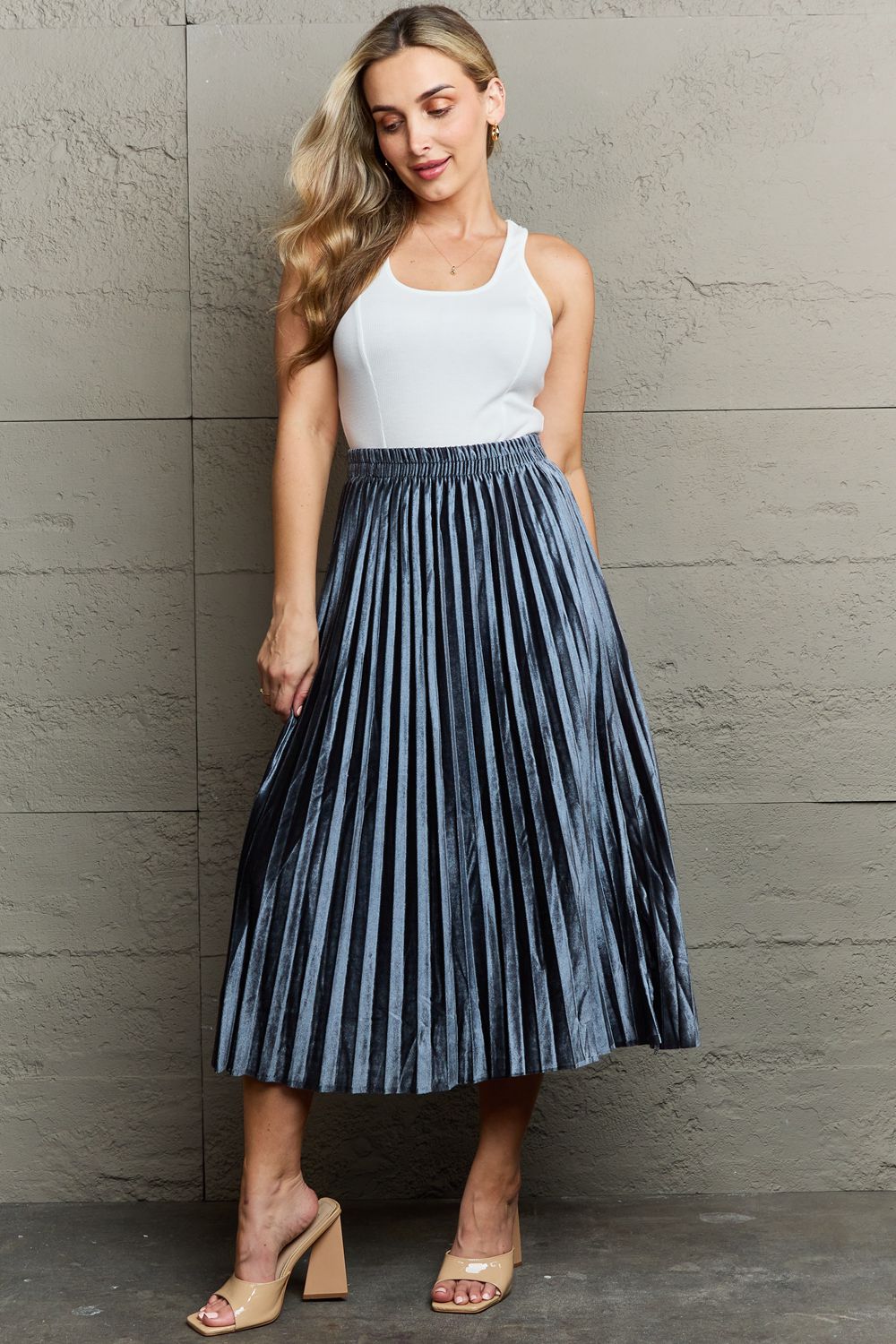 A person in a white top and the Ninexis Accordion Pleated Flowy Midi Skirt stands against a textured wall, wearing beige heeled sandals.