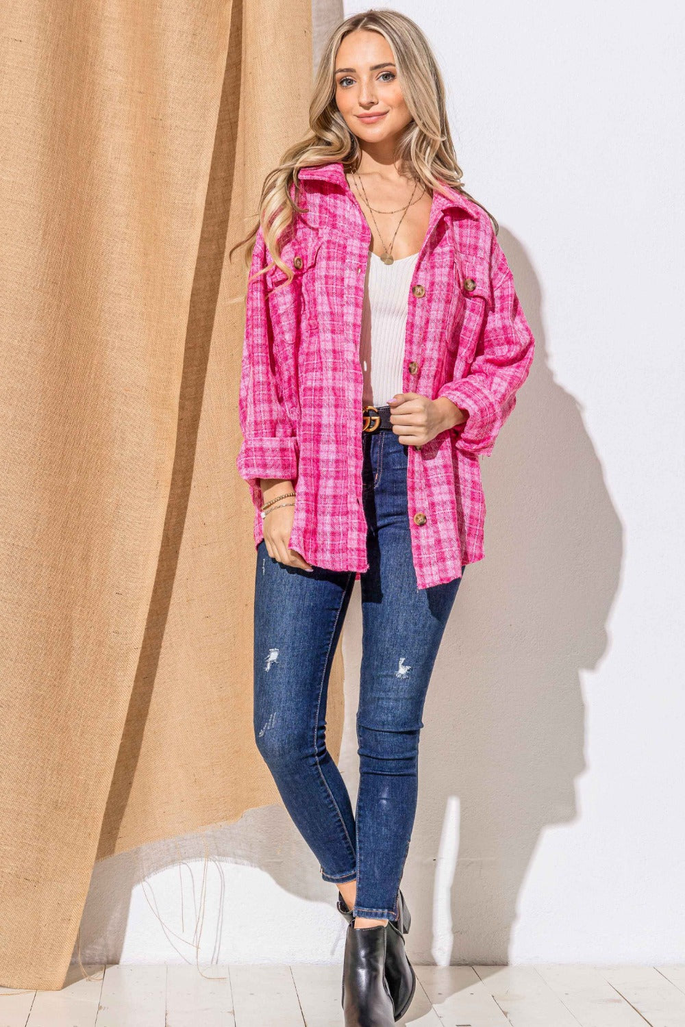 A woman stands smiling, wearing the "And The Why Full Size Tweed Button Down Long Sleeve Shacket" in pink plaid over a white top with a black belt and jeans. She is against a wall partially covered by a burlap curtain.