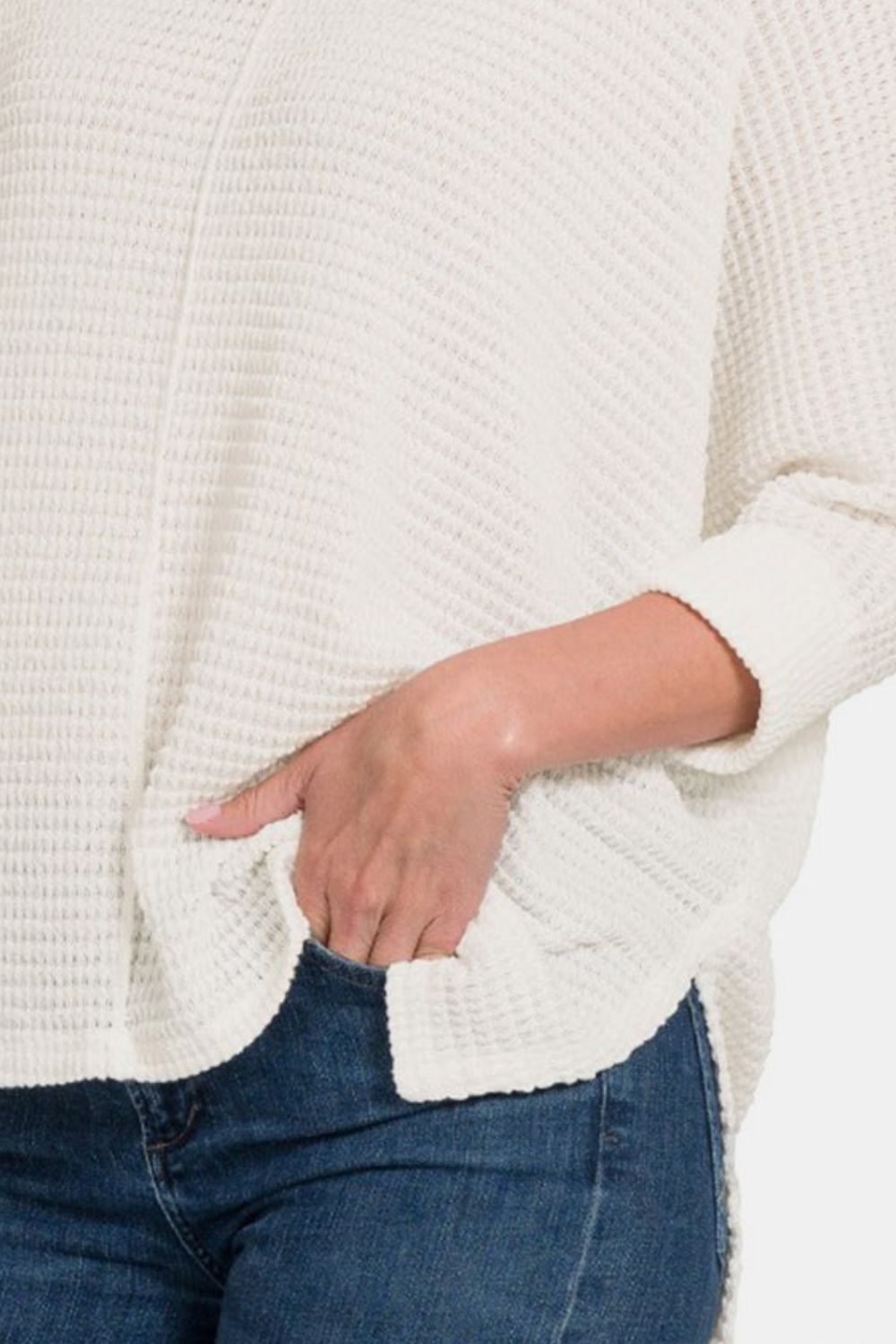 A smiling woman confidently poses in the Zenana Full Size Round Neck High-Low Slit Knit Top, which features a loose fit and three-quarter sleeves. She pairs the white knit top with blue jeans, striking a pose with one hand on her head and the other in her pocket.