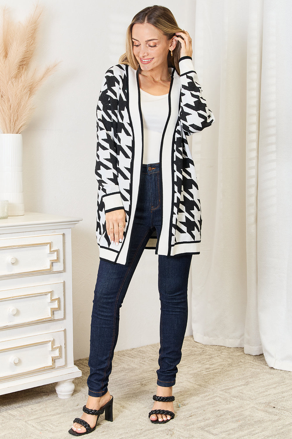 Woman wearing the Mandy Woven Right Houndstooth Open Front Longline Cardigan over a white top, standing next to a white chest of drawers with pampas grass in a vase on it.