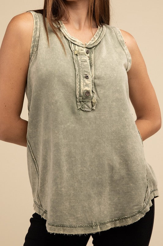 A woman stands against a beige background, wearing a Washed Half-Button Raw Edge Sleeveless Henley Top and black pants. She has long brown hair and holds the sides of her top.