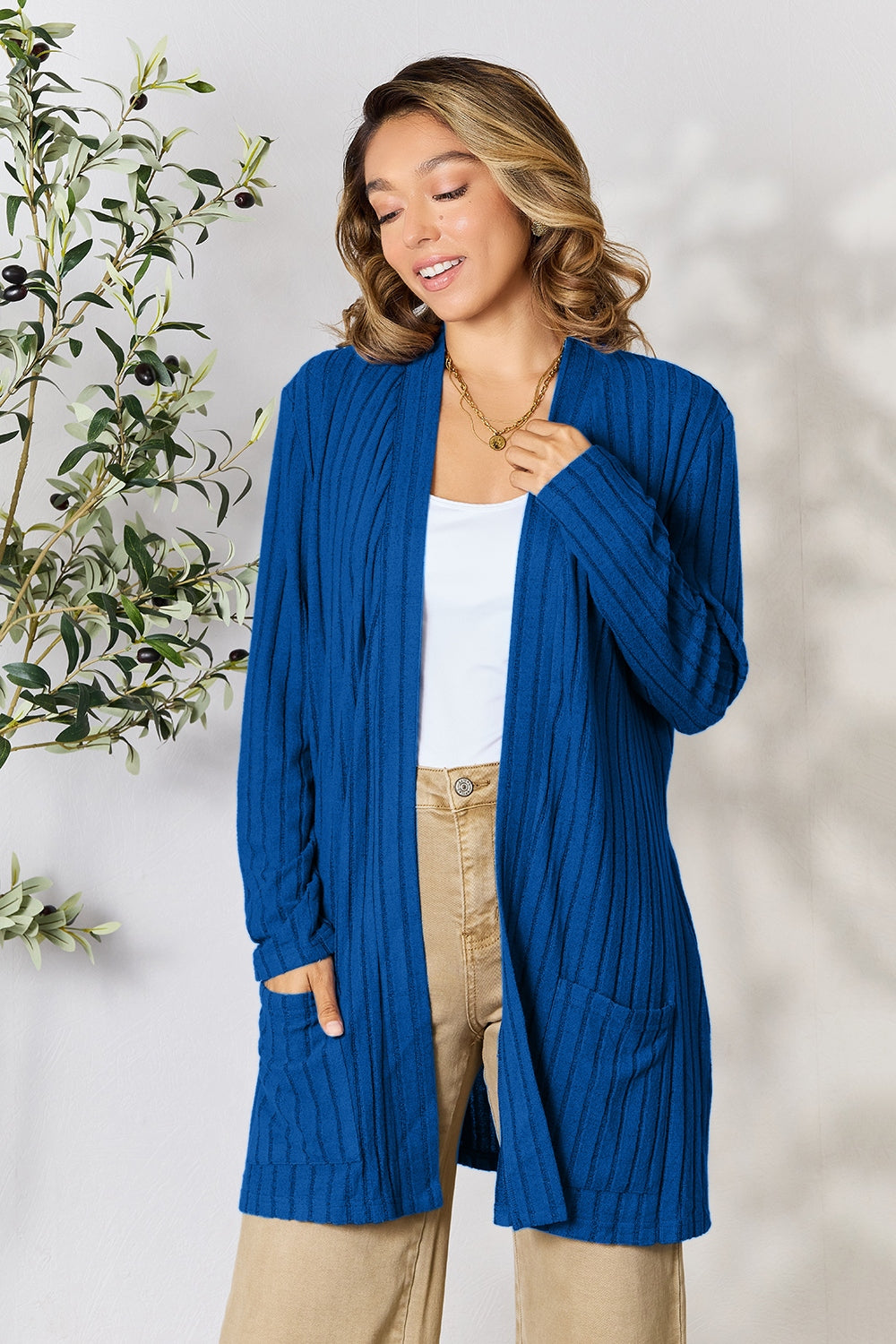 A woman in a Basic Bae Full Size Ribbed Open Front Cardigan with Pockets and jeans, made from stretchy material, stands in front of a plant, smiling slightly and looking to the side.