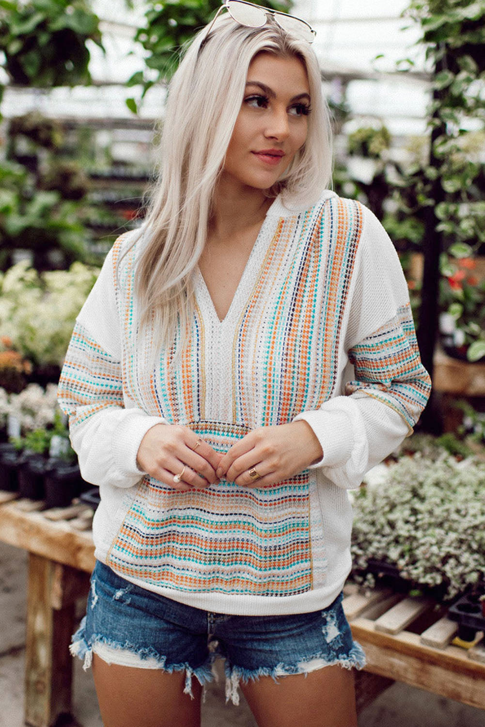 A woman with long blonde hair wearing a Multicolor Striped Patchwork V Neck Drop Shoulder Knit Hoodie and denim shorts stands in a plant nursery. She has sunglasses perched on her head and is looking down.