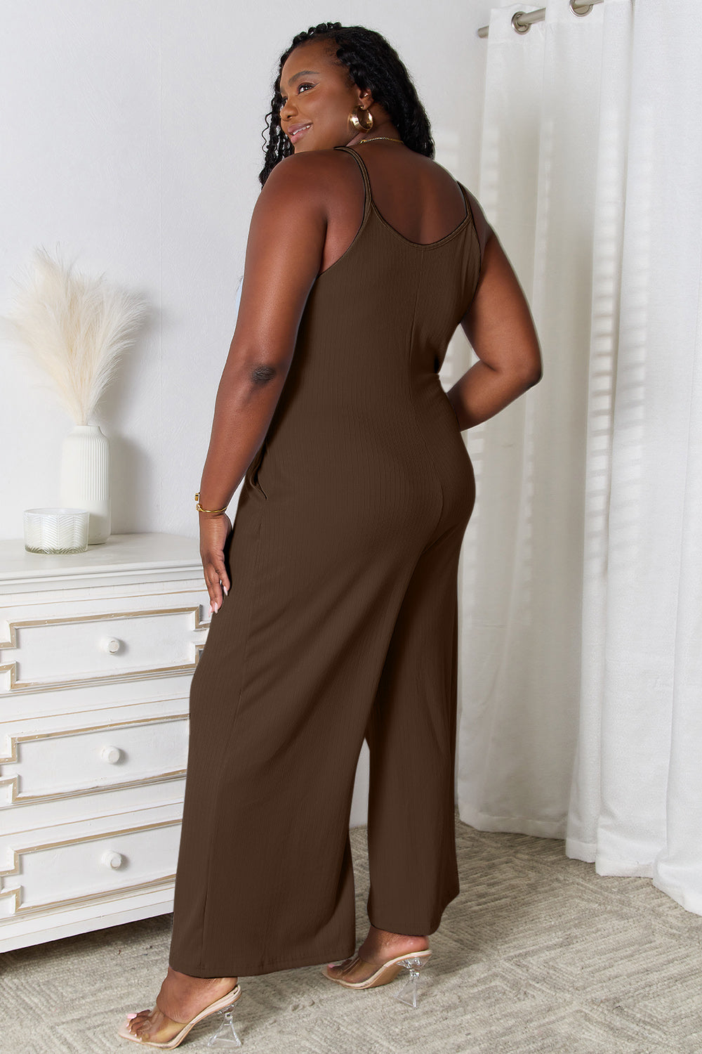 A woman in a stylish Basic Bae Full Size Spaghetti Strap V-Neck Jumpsuit stands indoors near a white dresser adorned with decorative items, smiling with one hand in her pocket.