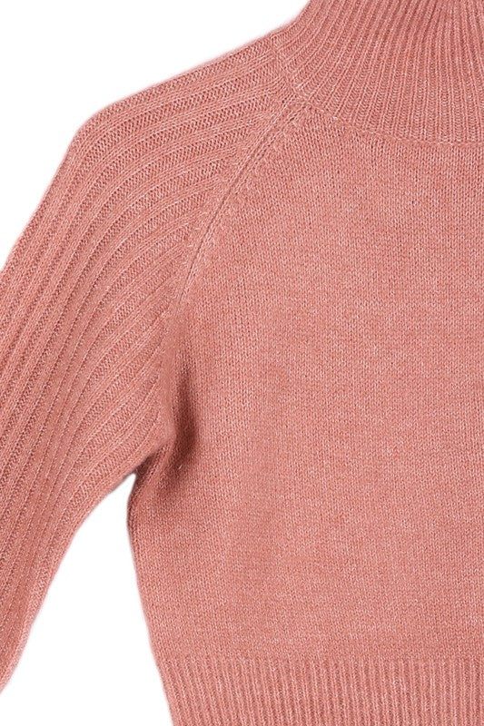 A person wearing a pink crop mock neck sweater with raglan sleeves poses with one hand near their chin.