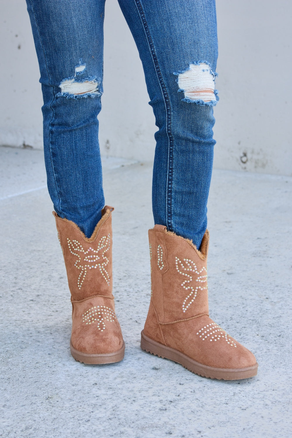 A person wearing ripped blue jeans and Forever Link Rhinestone Furry Flat Boots stands on a concrete surface.